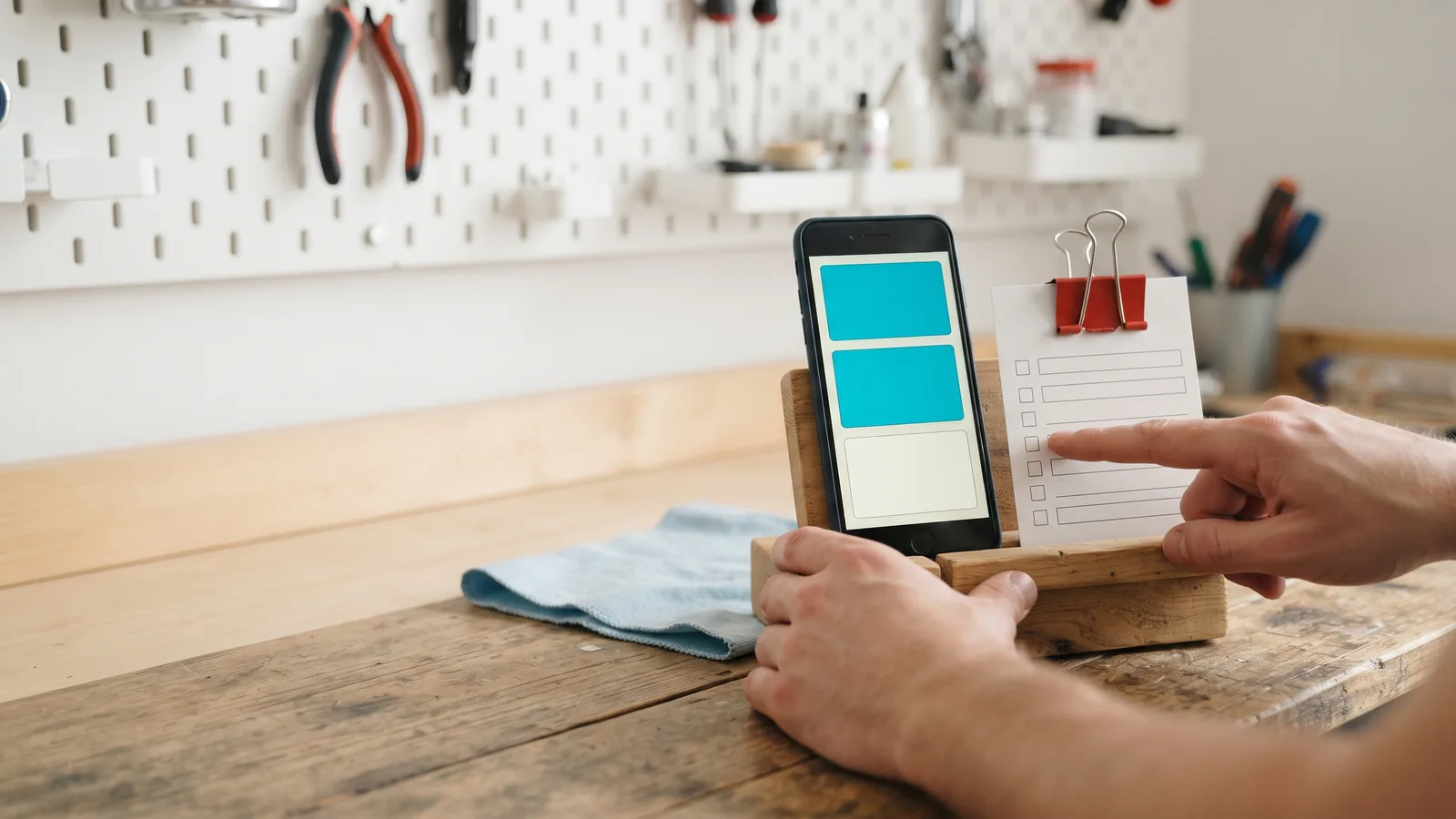 Hands point to a blank checklist with a red clip beside a phone in a wooden cradle on a workshop bench.