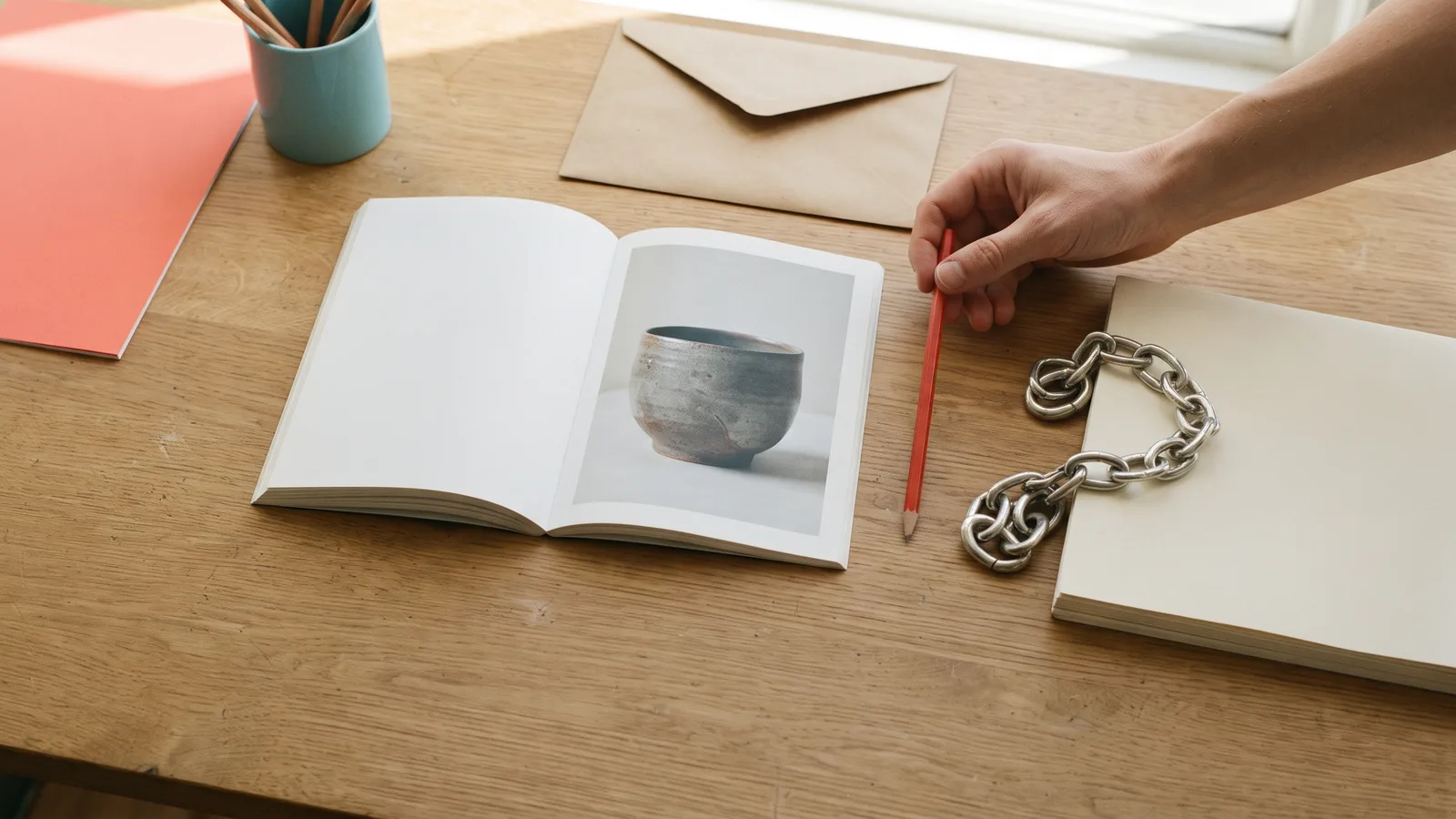 A hand holds a red pencil between an open pottery booklet and a metal chain on blank pages, beside kraft envelopes.
