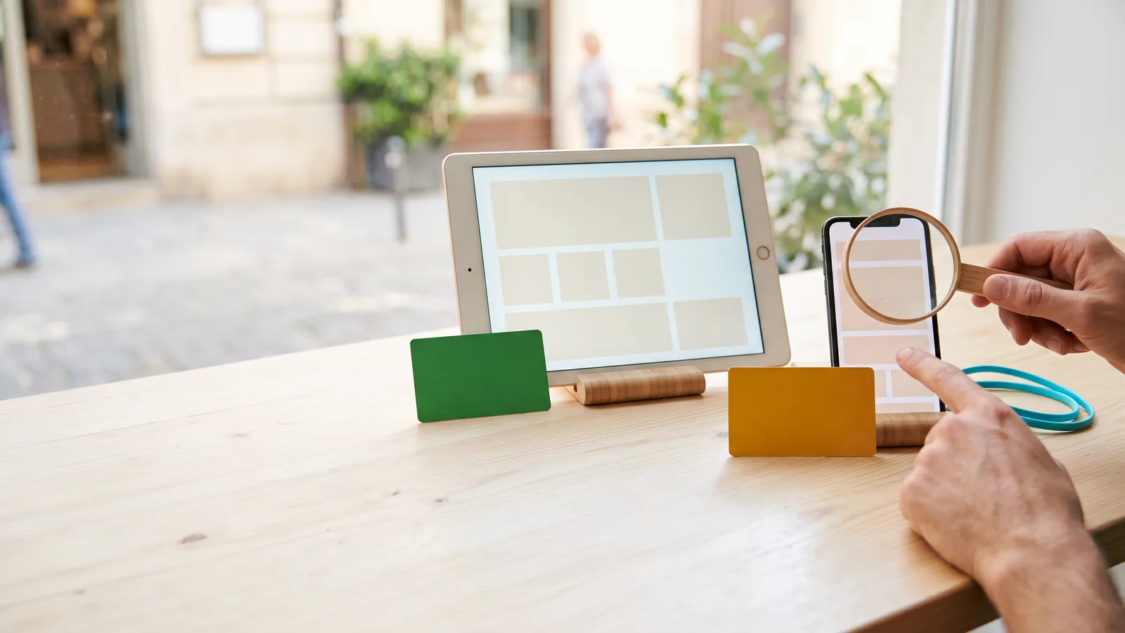 Hands use a magnifying glass to inspect a phone beside a tablet and coloured cards on a sunlit table.