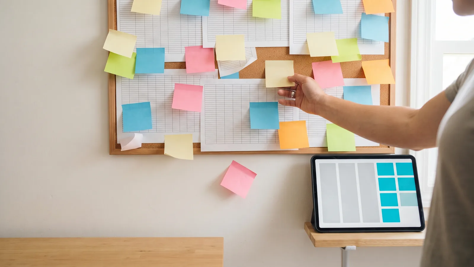A hand pins a sticky note on a cork board of grid sheets covered in notes; below, a tablet shows tidy columns of cyan blocks