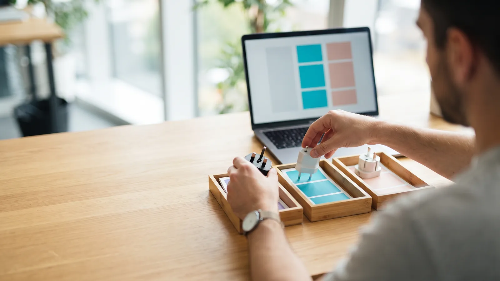 Hands sort British, European and Swiss plugs into three wooden trays of colour cards beside a laptop with matching blocks